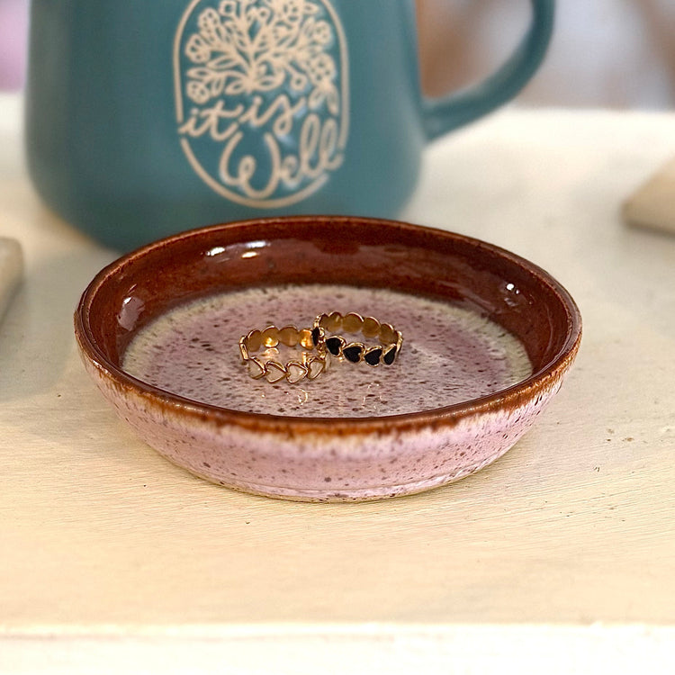 Jewelry dish with rings on a table next to a mug with 'it is well' text.